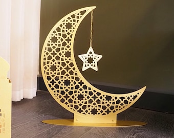 May include: A gold metal crescent moon with a star hanging from it. The moon is decorated with a geometric pattern. The star is also gold metal and has a simple design.