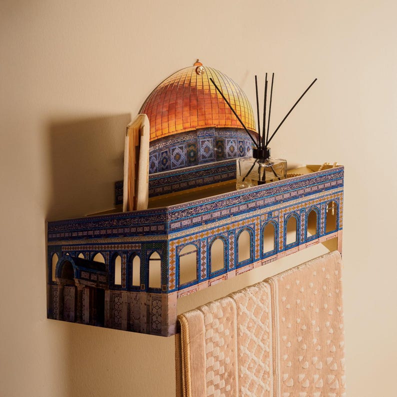 Pu&ograve; includere: Mensola decorativa con una dettagliata illustrazione della Cupola della Roccia, con una cupola dorata e dettagli architettonici blu e bianchi. La mensola contiene un libro, un diffusore e due asciugamani beige.