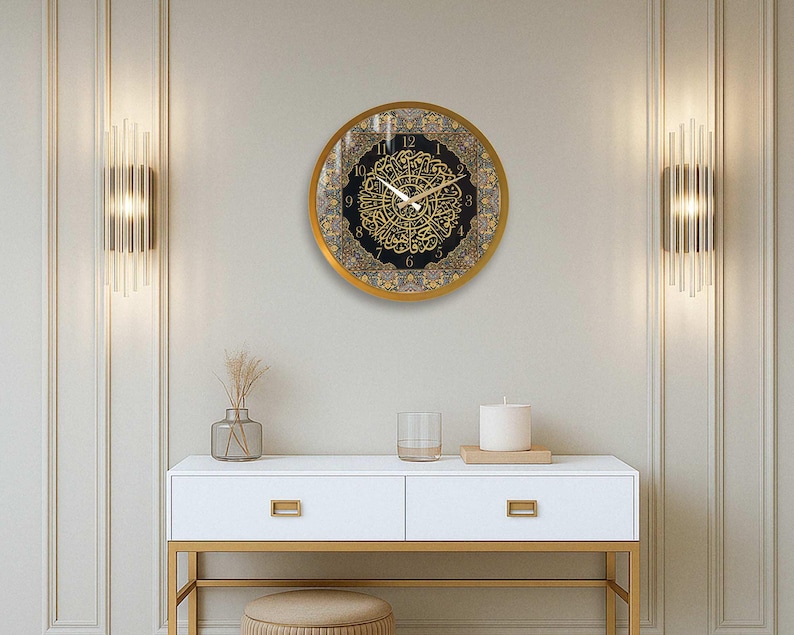 May include: A gold framed clock with black and gold Arabic numerals and a decorative design. The clock is hanging on a white wall with gold trim. A white table with gold legs is in the foreground.