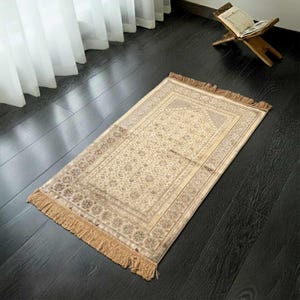 May include: A beige prayer rug with an intricate floral pattern and a fringed border. The rug is rectangular and lies on a dark wooden floor. A wooden book stand with an open book is in the background.