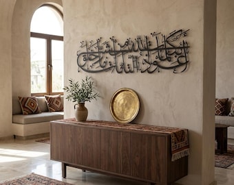 May include: A dark metal wall art piece featuring Arabic calligraphy hangs above a wooden cabinet. A gold-colored tray and a vase with greenery sit on the cabinet. The room has a neutral color palette with a rug and decorative pillows.