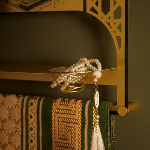 May include: A gold-colored metal wall shelf with an arched cutout design features a small circular platform holding a string of white pearl prayer beads. A green and tan patterned textile hangs from the lower bar of the shelf structure.