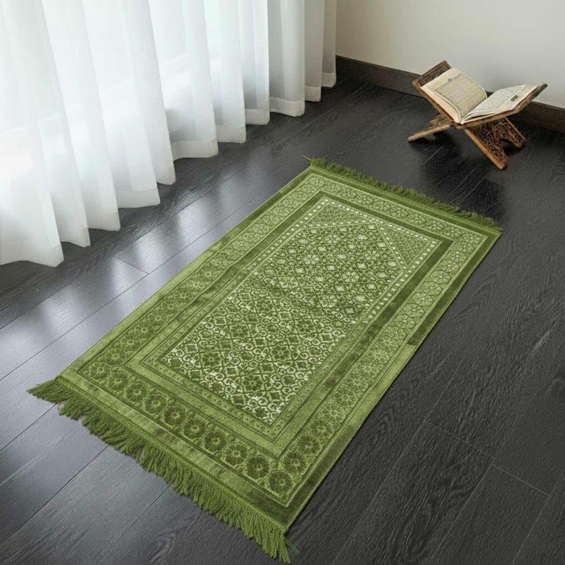 May include: A green prayer rug with an intricate floral pattern and fringe. The rug is rectangular and has a detailed border. A wooden book stand with an open book is in the background.