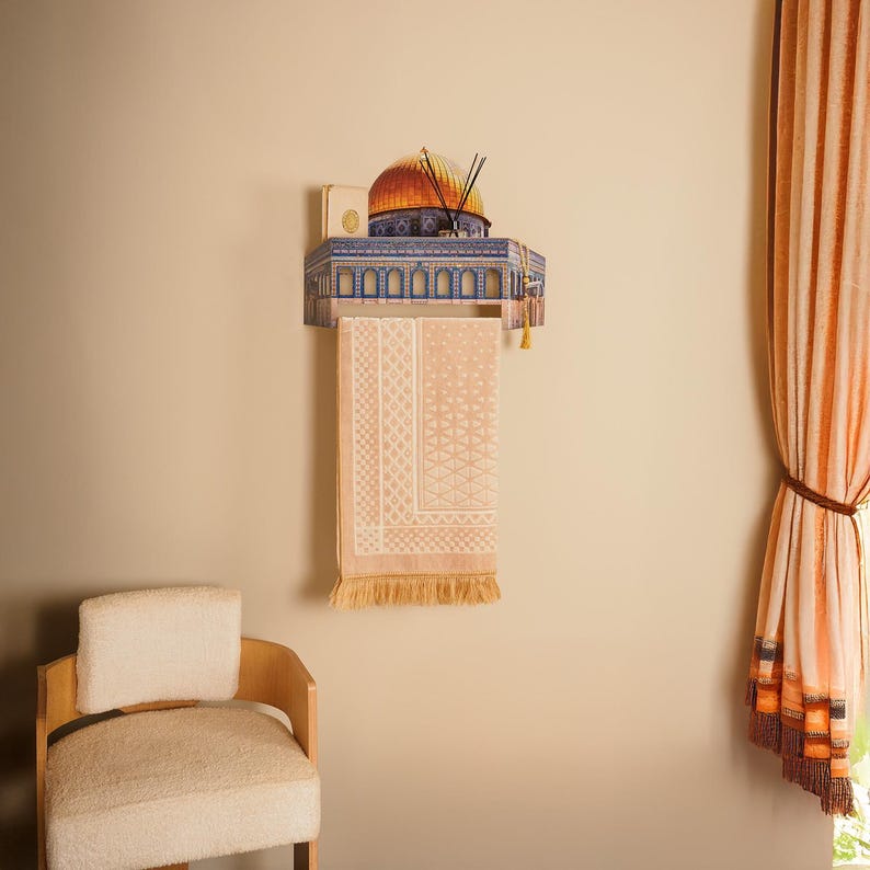 Pu&ograve; includere: Una mensola decorativa da parete con una dettagliata illustrazione di una struttura religiosa con una cupola dorata. Sotto, un tappeto da preghiera color pesca chiaro con motivi geometrici &egrave; appeso. La mensola contiene bastoncini di incenso e un piccolo libro.