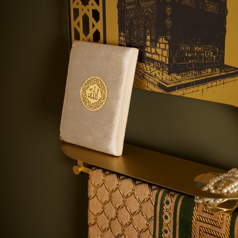 May include: A beige velvet-covered book featuring a circular gold emblem with Arabic calligraphy centered on the cover. The book rests on a gold-toned metal shelf, which also holds a patterned textile with green and tan geometric designs.