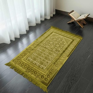 May include: A green prayer rug with a detailed floral pattern and fringe. The rug is rectangular and lies on a dark wooden floor. A wooden book stand with an open book is in the background.