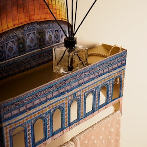 Pu&ograve; includere: Una mensola decorativa con un design architettonico mediorientale. La mensola presenta un interno dorato, un esterno blu e oro e un flacone diffusore in vetro trasparente con canne nere. Una collana di perle poggia sulla mensola.