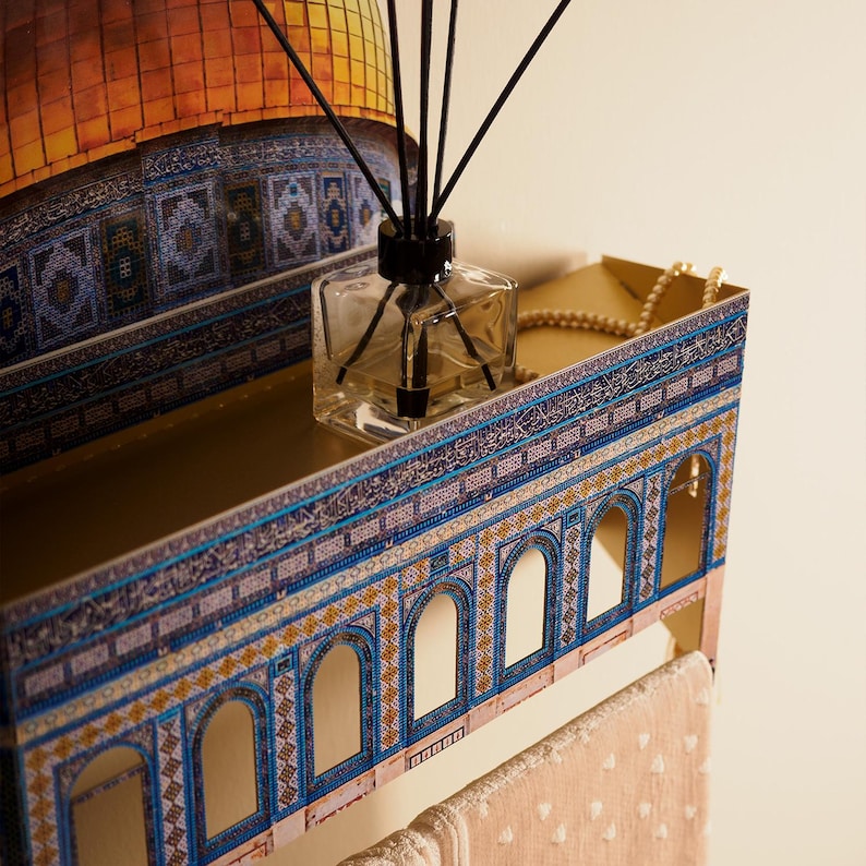 Pu&ograve; includere: Una mensola decorativa con un design architettonico mediorientale. La mensola presenta un interno dorato, un esterno blu e oro e un flacone diffusore in vetro trasparente con canne nere. Una collana di perle poggia sulla mensola.