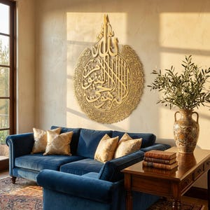 May include: A gold-colored, circular wall art piece with Arabic calligraphy hangs above a blue velvet sofa with gold patterned pillows. A wooden side table holds books and a decorative vase with greenery.