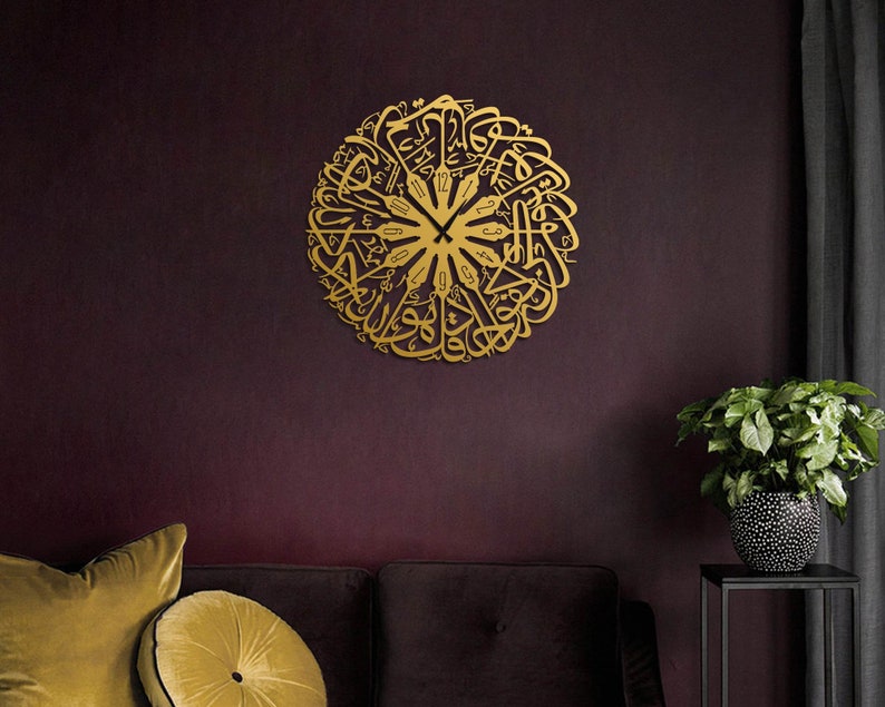 May include: A gold-colored, circular wall clock with Arabic calligraphy. The clock is mounted on a dark burgundy wall, with a sofa and decorative pillows in the foreground. A potted plant sits to the right.