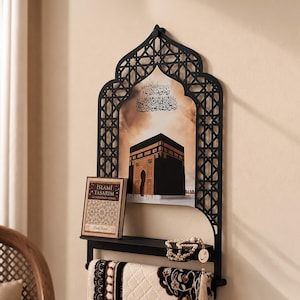 May include: A black metal wall shelf featuring an arched, laser-cut geometric frame. The center displays an image of the Kaaba with Arabic calligraphy. A small book titled ISLAMI TASARIM, a string of prayer beads, and a prayer rug are included.