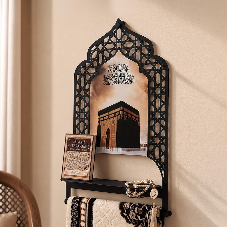 May include: A black metal wall shelf featuring an arched, laser-cut geometric frame. The center displays an image of the Kaaba with Arabic calligraphy. A small book titled ISLAMI TASARIM, a string of prayer beads, and a prayer rug are included.
