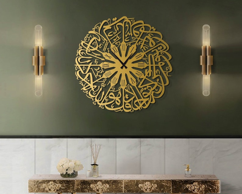 May include: A gold-colored, circular wall clock featuring Arabic calligraphy. The clock has black hands and Roman numerals at the top. The clock is mounted on a dark green wall, with two gold sconces on either side. Below the clock is a marble-topped console.