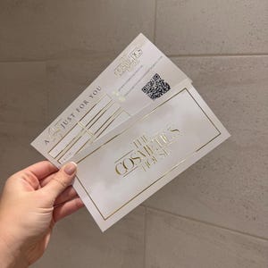 May include: Two white gift cards with gold lettering and accents are held in a hand. The cards read "The Cosmetics House" and "Just For You". A QR code is visible on one card.