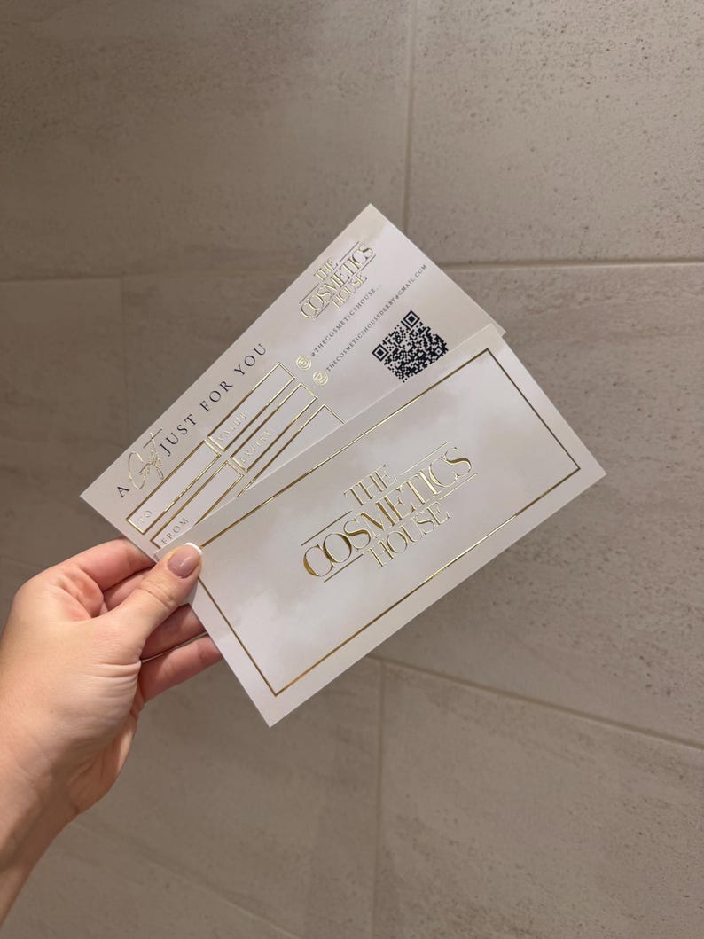 May include: Two white gift cards with gold lettering and accents are held in a hand. The cards read "The Cosmetics House" and "Just For You". A QR code is visible on one card.