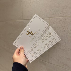 May include: Two white business cards with gold foil accents are held in a hand. The cards feature the text "SOPHIE ELIZABETH AESTHETICS" and a logo. One card has lines for information.