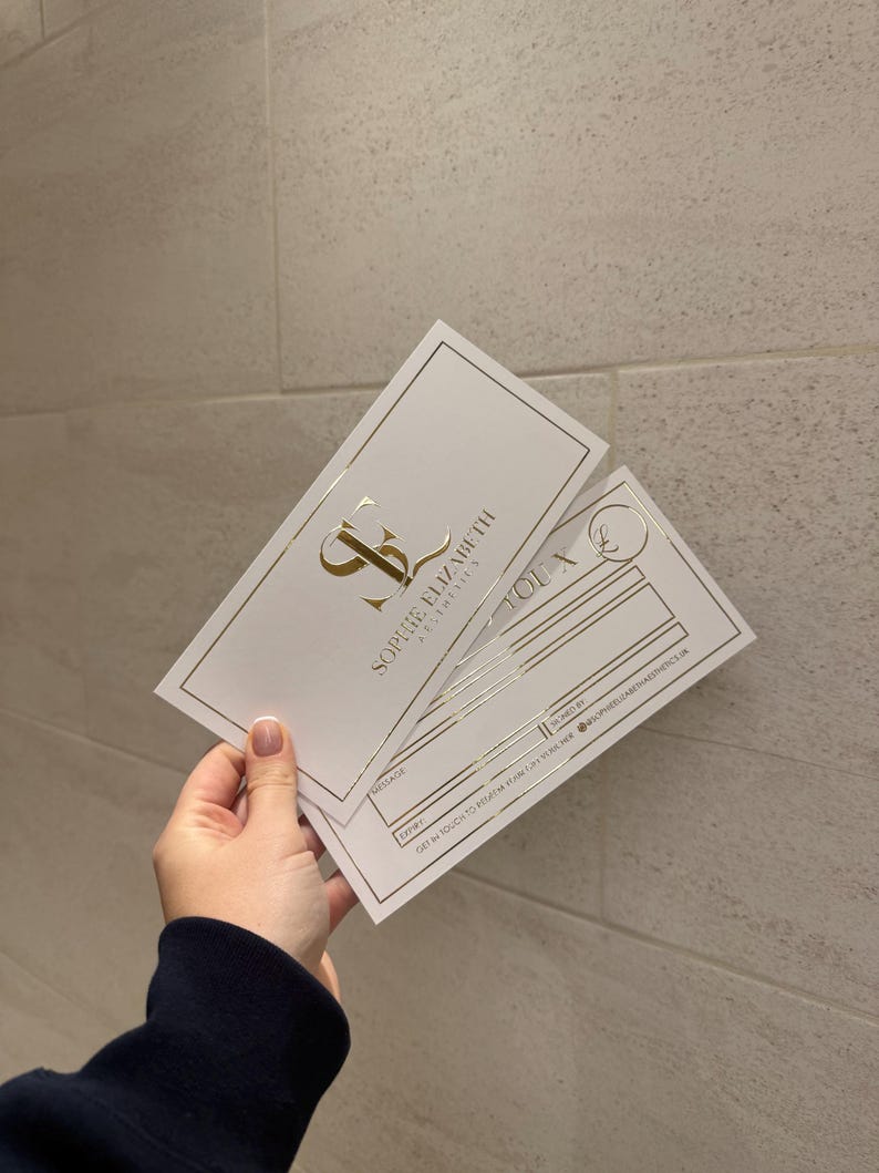 May include: Two white business cards with gold foil accents are held in a hand. The cards feature the text "SOPHIE ELIZABETH AESTHETICS" and a logo. One card has lines for information.