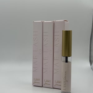 May include: Three pink boxes and a clear tube of lash serum. The boxes are labelled "MAE LASH CO" and "LASH Jeran". The tube has a gold cap and the words "LASH Serum" printed on it. The background is a plain white.