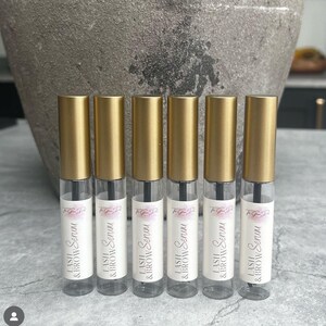 以下が含まれることがあります： 黒いキャップと白いラベルに「LASH & BROW Serum」と書かれた、金色と透明なボトルが6本あります。