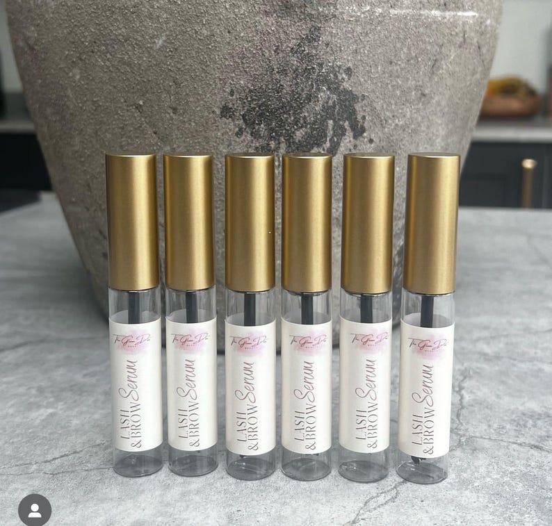 以下が含まれることがあります： 黒いキャップと白いラベルに「LASH & BROW Serum」と書かれた、金色と透明なボトルが6本あります。