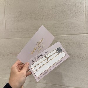 May include: A gift certificate from Medi-Refine BYG Aesthetics. The pink card features gold text and a QR code. The card reads "A Gift For You" with spaces for recipient, sender, value, and a message. The business logo and contact information are also visible.