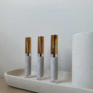 以下が含まれることがあります： 白いテクスチャ加工された表面に、金色の蓋と「LASH OIL」と書かれたラベルが付いた透明なガラス瓶が3本あります。
