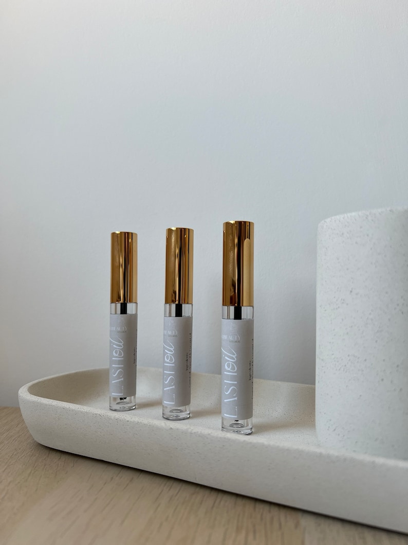 以下が含まれることがあります： 白いテクスチャ加工された表面に、金色の蓋と「LASH OIL」と書かれたラベルが付いた透明なガラス瓶が3本あります。