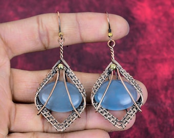 May include: A pair of dangle earrings featuring oval-shaped, translucent light blue stones encased in intricate, hand-woven copper wire frames. Each earring is suspended from a simple copper hook and accented with a small, polished gold-tone bead.