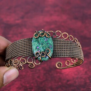 May include: A handcrafted copper cuff bracelet with a central oval green stone with red flecks. The bracelet has a woven wire design and decorative copper swirls and loops.