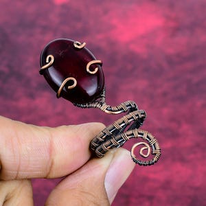 Puede incluir: Anillo artesanal con una piedra preciosa ovalada de color rojo oscuro. La piedra está engastada en un diseño de alambre de color cobre que se enrolla alrededor del dedo. El anillo tiene un aspecto artístico único.