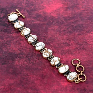 May include: A gold-toned bracelet with oval white and brown stones. The bracelet has a toggle clasp and a series of three connected rings at the end. The stones are set in a dark metal frame.