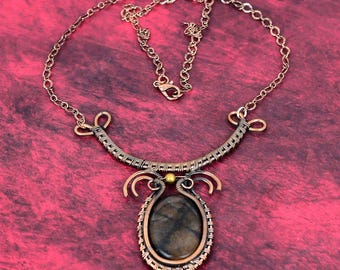 May include: A wire-wrapped necklace featuring a central oval dark brown stone with a cross pattern, encased in copper wire. The pendant connects to a curved copper wire collar with decorative loops and a small gold bead, attached to a copper chain.