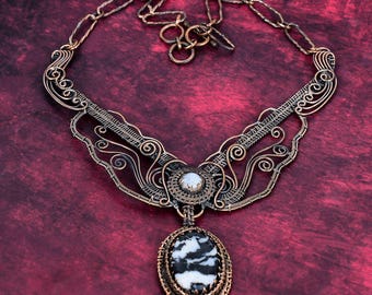 May include: A copper wire-wrapped necklace featuring an oval black and white patterned stone pendant, a smaller round white stone accent, and intricate metal scrollwork. The piece includes a chain with oval links and a toggle clasp closure.