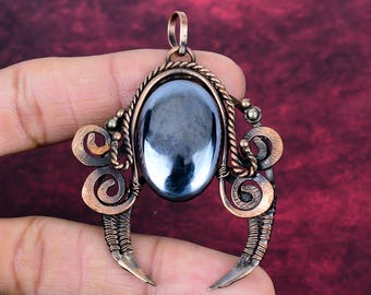 May include: A copper wire-wrapped pendant featuring a central oval-shaped, polished dark gray hematite stone. The metal frame is constructed with twisted wire accents, decorative spirals on both sides, and two downward-curving points.