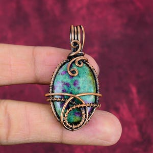 May include: Oval pendant featuring a green and purple stone, intricately wrapped in copper wire. The wirework includes decorative swirls and a bail for a necklace. The pendant is held against a burgundy background.