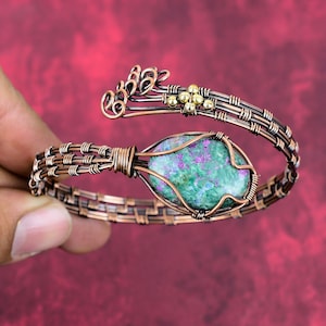 May include: A handcrafted copper wire bracelet with a large, oval-shaped, green and purple stone at its center. The bracelet features intricate wirework and small gold-colored accents, creating a unique and artistic design. The bracelet is shown against a red background.