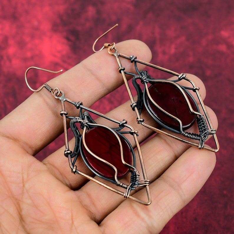 May include: A pair of dangle earrings with a geometric diamond shape. The earrings are made of copper wire and feature a red gemstone in the center.
