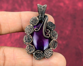 May include: An oval-shaped purple gemstone pendant encased in dark copper wire. The wire is intricately coiled into a series of small, circular rose-like patterns surrounding the stone, with a decorative wire bail at the top for suspension.