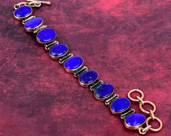 Könnte beinhalten: Ein goldfarbenes Armband mit ovalen, tiefblauen Edelsteinen. Das Armband hat einen Knebelverschluss und liegt auf einem weinroten Hintergrund. Die Edelsteine sind gleichmäßig angeordnet und von der Goldfassung umrahmt.