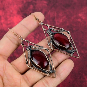 May include: A pair of dangle earrings with a geometric design. The earrings are made of copper wire and feature a red gemstone in the center of each earring.