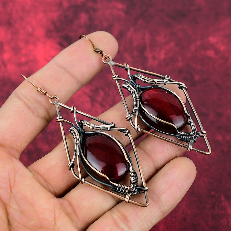 May include: A pair of dangle earrings with a geometric design. The earrings are made of copper wire and feature a red gemstone in the center of each earring.