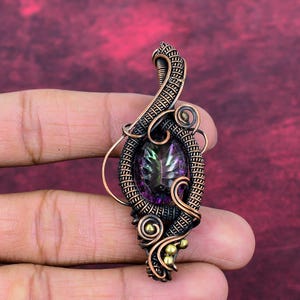 Peut inclure: Un pendentif artisanal avec une pierre précieuse irisée dans les tons de violet, vert et or. La pierre est sertie dans un fil de cuivre complexe, avec des spirales décoratives et de petits accents en laiton. Le pendentif est tenu dans une main.
