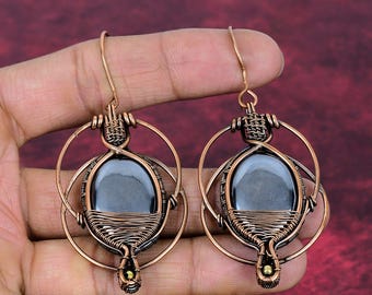 May include: A pair of wire-wrapped earrings featuring oval-shaped, dark gray, metallic-looking stones encased in copper wire. The design includes a circular copper frame surrounding the central stone, with a hook-style closure at the top.