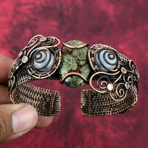 May include: Copper wire cuff bracelet featuring a central oval green stone with red and brown markings, flanked by two round, light-colored stones. The bracelet has intricate wirework with spiral and woven patterns.