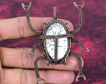 Peut inclure: Pendentif en fil de cuivre en forme de scarabée. Il présente une pierre ovale blanche veinée de noir, sertie dans une monture en fil de cuivre. Le scarabée a six pattes et deux antennes, toutes en fil de cuivre. Le bijou est tenu dans une main.