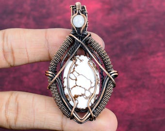 May include: A wire-wrapped pendant featuring a central oval white stone with brown veining, encased in oxidized copper wire. A small, round, translucent white gemstone is set at the top bail, integrated into the intricate copper wirework design.