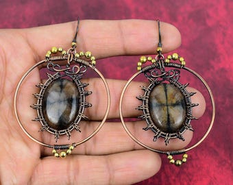 May include: A pair of handcrafted earrings featuring large, oval-shaped stones with brown and gray hues. The stones are set within intricate, dark metal wirework, surrounded by a copper-colored hoop and accented with gold-colored beads. The earrings are displayed against a red background.