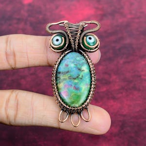 Peut inclure: Un pendentif hibou artisanal en fil de cuivre avec un grand corps en pierre ovale verte et rouge. Le hibou a deux perles vertes pour les yeux et des détails en fil de cuivre. Le pendentif est tenu sur un fond bordeaux.