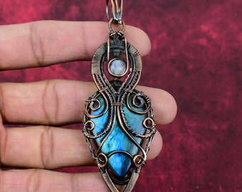 Peut inclure: Un pendentif en fil de cuivre enroulé avec une pierre précieuse en labradorite bleue et une plus petite pierre précieuse blanche. Le pendentif a la forme d'une larme et présente un travail de fil complexe.