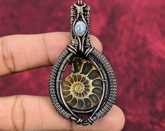 Puede incluir: Colgante artesanal con un amonita fosilizada encapsulada en alambre de cobre. El colgante tiene un diseño en espiral con una pequeña piedra redonda de color azul grisáceo en la parte superior. El trabajo de alambre está intrincadamente tejido, creando una estética única y terrosa.